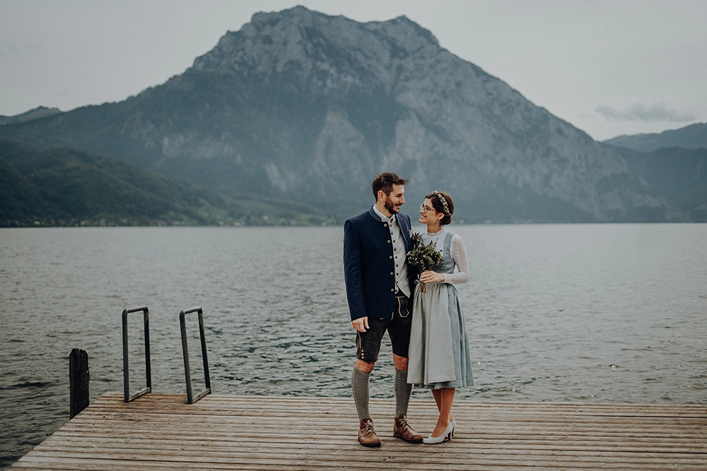 Brautpaar in Tracht steht auf einem Steg. See und Berg im Hintergrund